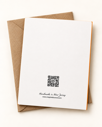 A white stationery card with a black QR code and small printed text at the bottom, placed on top of a brown kraft envelope against a light beige background.