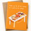 An orange Thanksgiving greeting card with a brown envelope behind it. The card features the text 'Thank you for all the cooking so I could do the eating!' above an illustration of a Thanksgiving table filled with food, and 'HAPPY THANKSGIVING' written near the bottom.