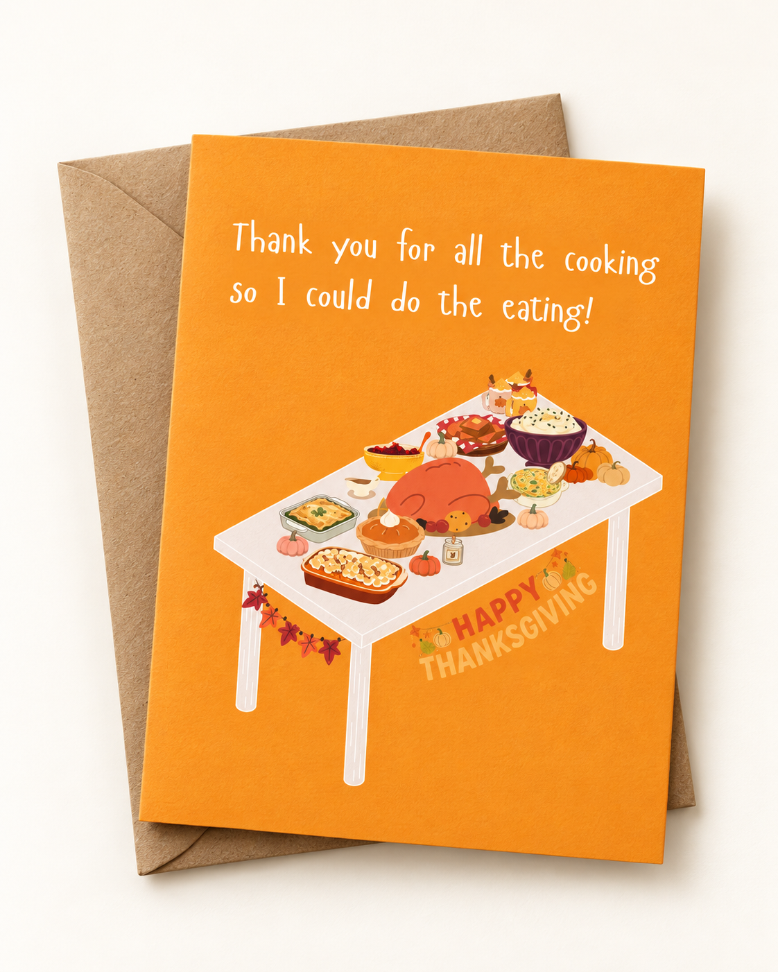 An orange Thanksgiving greeting card with a brown envelope behind it. The card features the text 'Thank you for all the cooking so I could do the eating!' above an illustration of a Thanksgiving table filled with food, and 'HAPPY THANKSGIVING' written near the bottom.