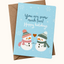 A square holiday greeting card with a light blue background, featuring two cartoon snowmen in winter hats and scarves, plus the text 'You are snow much fun! Happy holidays.' The card is shown with a brown envelope behind it on a light cream background.