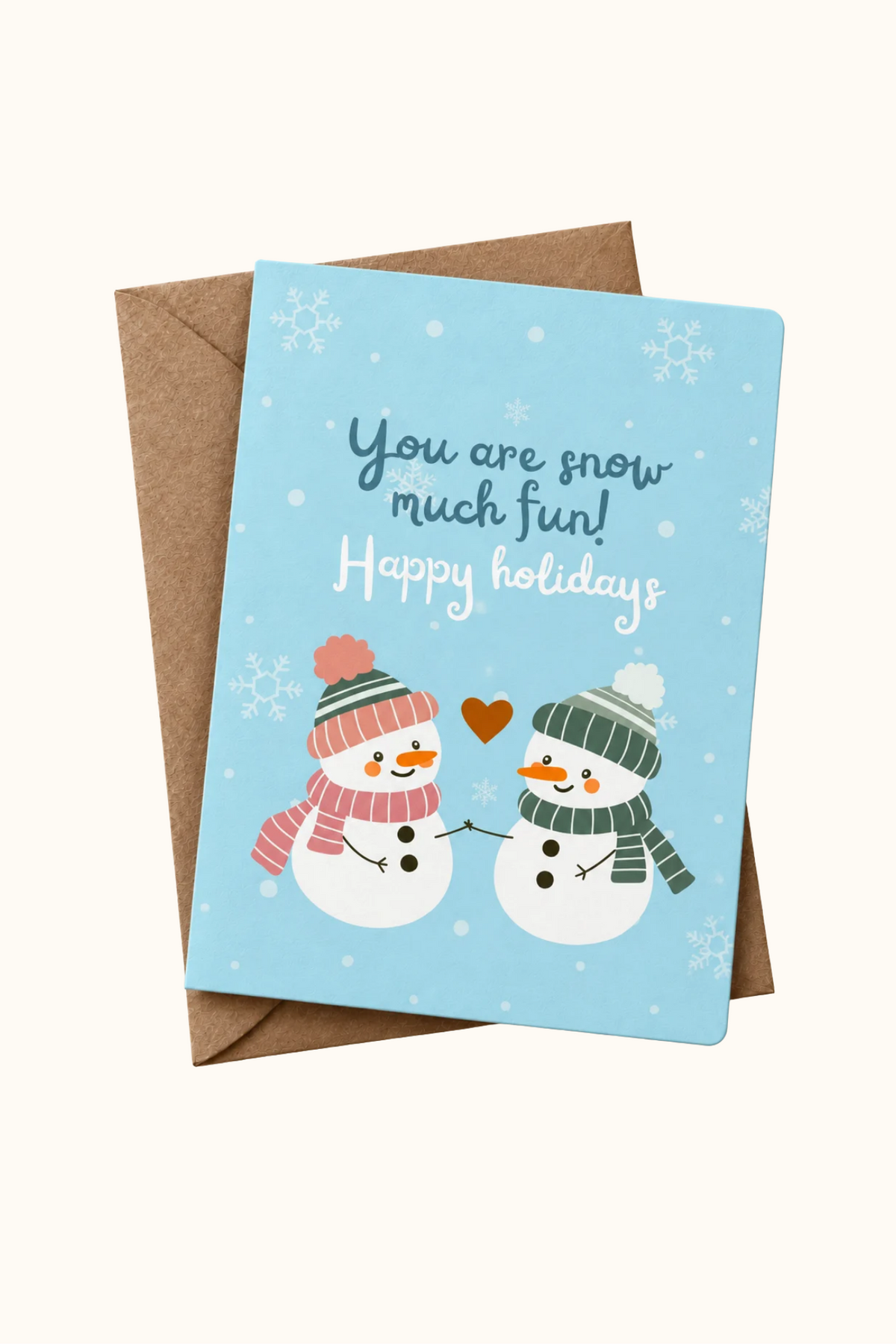 A square holiday greeting card with a light blue background, featuring two cartoon snowmen in winter hats and scarves, plus the text 'You are snow much fun! Happy holidays.' The card is shown with a brown envelope behind it on a light cream background.